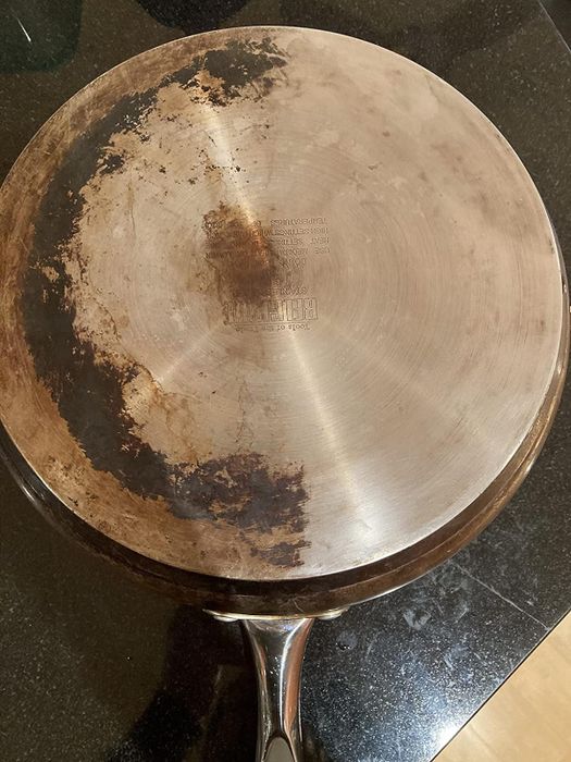 Well-used stainless steel frying pan on a stovetop, with one half shiny and clean, and the other half covered in stains