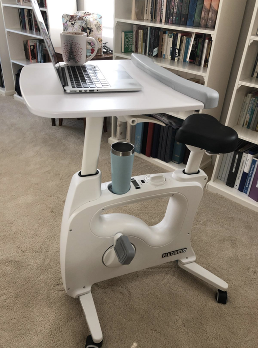 Reviewer image of a sleek white desk bike in a home office, with a laptop resting on the tray and a drink conveniently placed in the holder