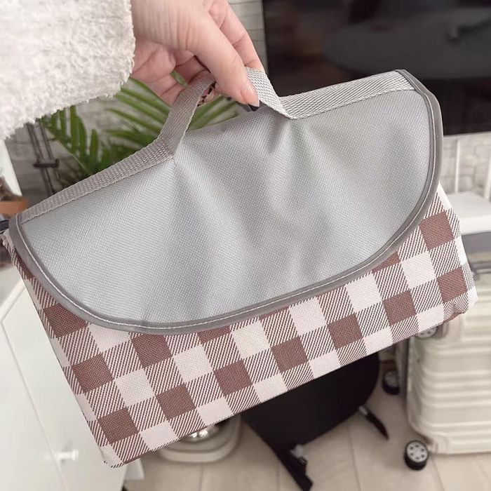 A hand holding a gray and brown plaid foldable picnic blanket with a handle, perfect for outdoor shopping or travel