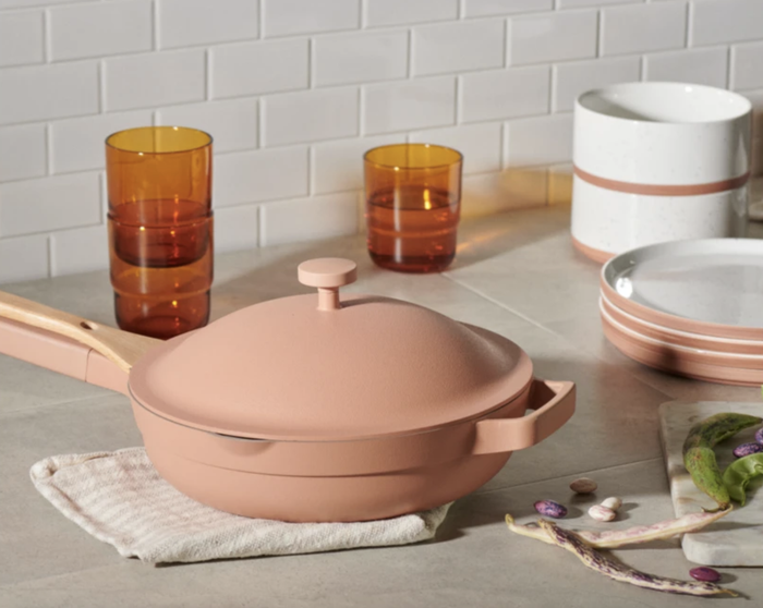 A pale peach Always Pan rests on a kitchen counter, surrounded by amber-colored cups and peach-toned dishes.