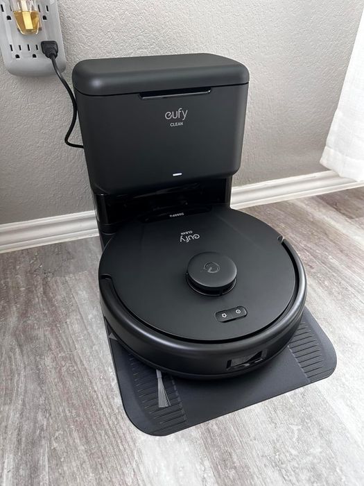 The Eufy RoboVac G40+, an automatic vacuum, is pictured resting in its charging station against a wall on a wooden floor with a power outlet in the background.