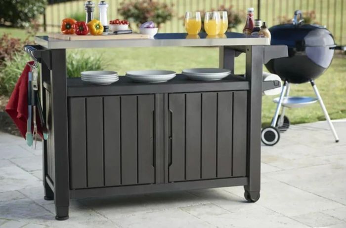 A dark buffet table with a metal top, stocked with ingredients and dishware, placed in front of a grill.