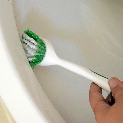 A hand using a scrub brush to clean the area under the rim of a toilet.