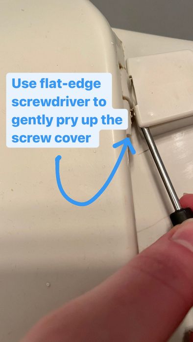 A person using a flat-head screwdriver to lift the screw cover on the back of a toilet seat.