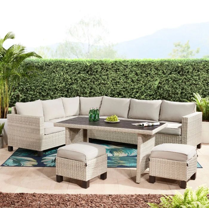A tan wicker sectional, dining table, and ottomans with plush tan cushions arranged in a beautifully decorated patio space.
