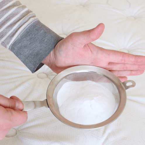 A blogger's hands using a kitchen sifter to sprinkle baking soda over a mattress.