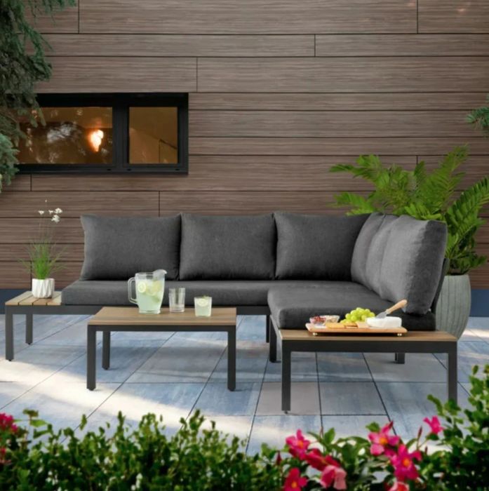 A grey sectional with light wood tables attached on both ends, complemented by a separate light wood coffee table, set in a charming patio environment.
