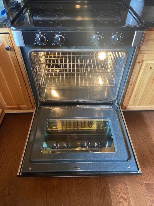 Open oven with the door down and the light on, revealing an empty interior, set in a wooden kitchen space. The floating electric stovetop is positioned above the oven.