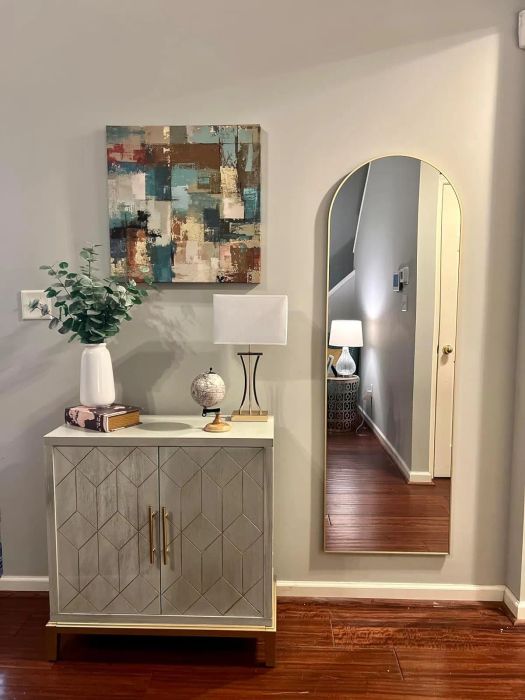 A stylish living room arrangement featuring a geometric cabinet, a potted plant, a globe, a lamp, an abstract wall painting, and an arched mirror reflecting a lamp and a staircase.
