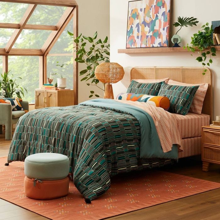 A cozy bedroom featuring patterned bedding, modern decor, and lush potted plants. The large window lets in ample natural light, enhancing the serene and peaceful atmosphere.