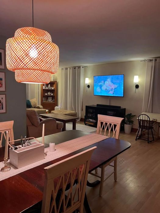 A modern dining area featuring a wooden table, chairs, an elegant ceiling light fixture, and a cozy living space with a TV and furniture in the background.