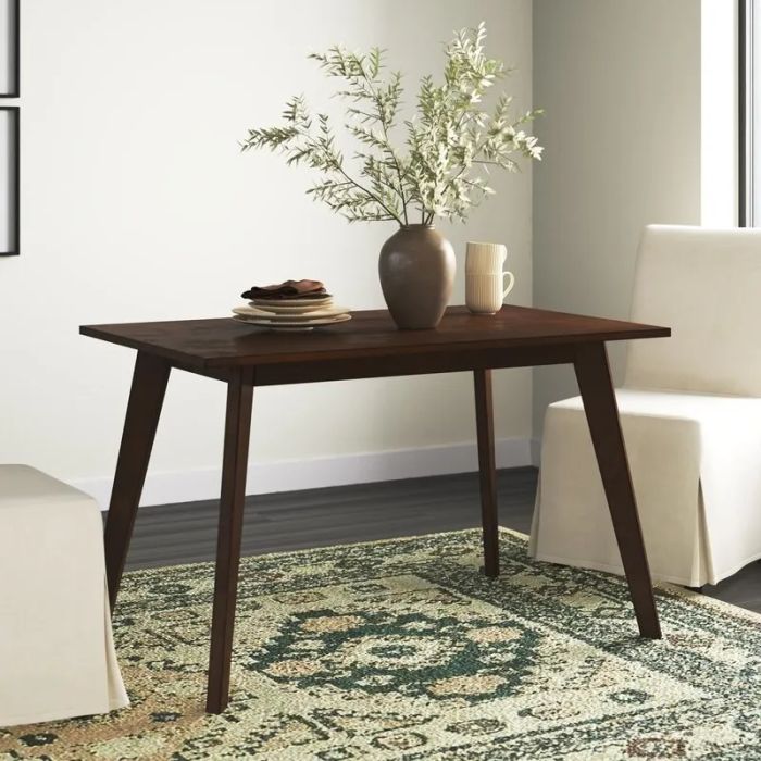 A modern wooden dining table with a vase and cup atop it, surrounded by white chairs on a patterned rug, in a bright and airy room.
