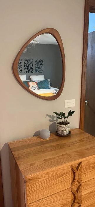 A wood-framed triangular mirror hangs above a wooden dresser, accompanied by a potted plant and a round decorative object, reflecting a bedroom with a tree-patterned headboard.