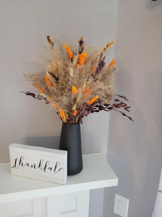 A black vase filled with a dried floral arrangement sits next to a white wooden sign that reads,