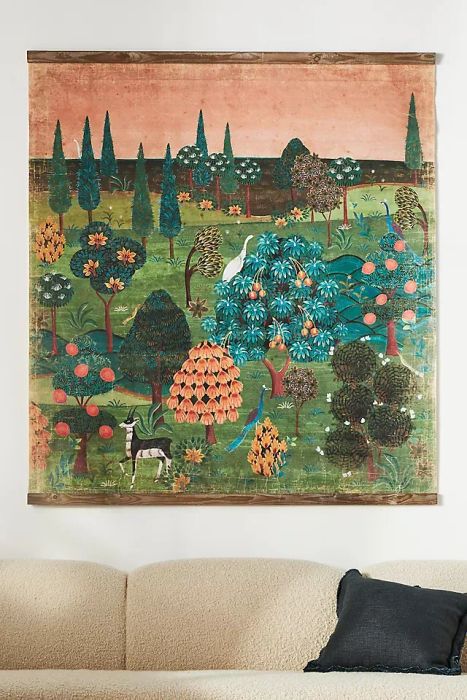 A large decorative tapestry featuring a vibrant landscape with trees, shrubs, a bird, and a deer, hanging above a white sofa adorned with a black pillow.