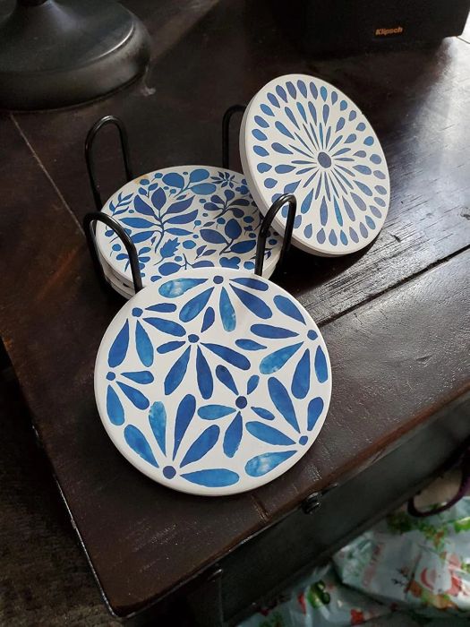 A set of four decorative coasters featuring blue floral and geometric designs, neatly arranged in a black metal holder atop a dark wooden table.