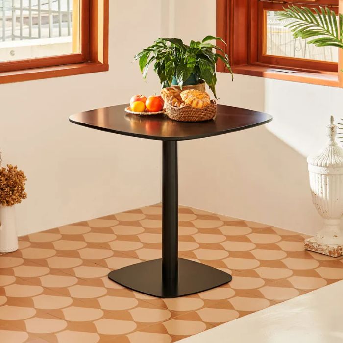 A contemporary square dining table with a pedestal base, featuring a plant, a basket of bread, and a bowl of fruit, positioned in a bright and airy room.