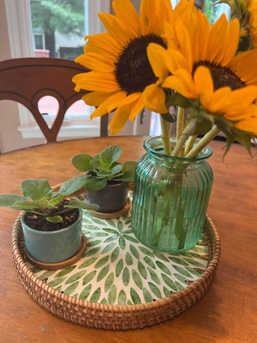 A set of sunflowers in a textured green glass vase, with two small potted plants arranged on a leafy-patterned tray resting on a wooden table.
