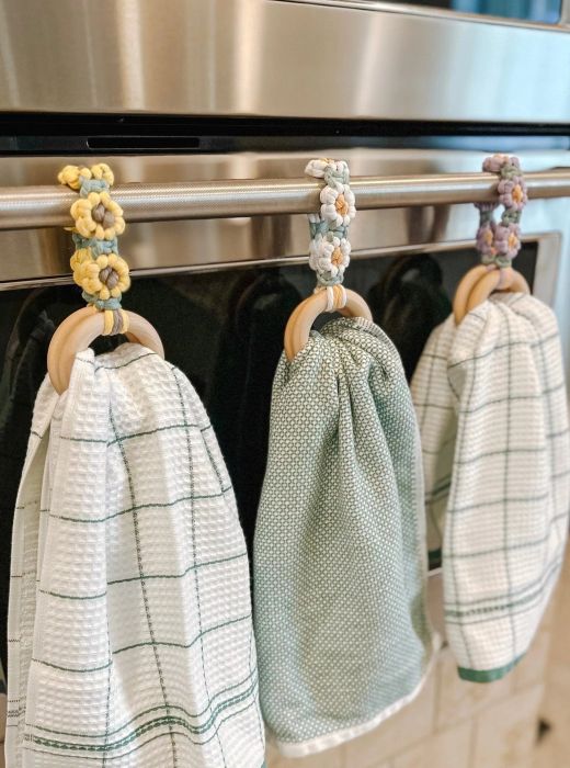 Three kitchen towels hanging from a stainless steel oven handle, each neatly secured with crocheted loop holders.