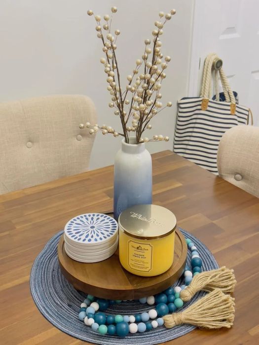 A styled table featuring a vase of branches, a yellow candle, blue and white coasters, and a blue beaded tassel. In the background, a striped bag hangs from a door handle.