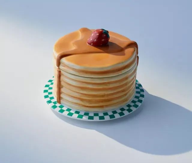 A stack of pancakes topped with syrup and a strawberry, served on a plate with a green-and-white checkered rim. AI-generated image for a shopping guide article.