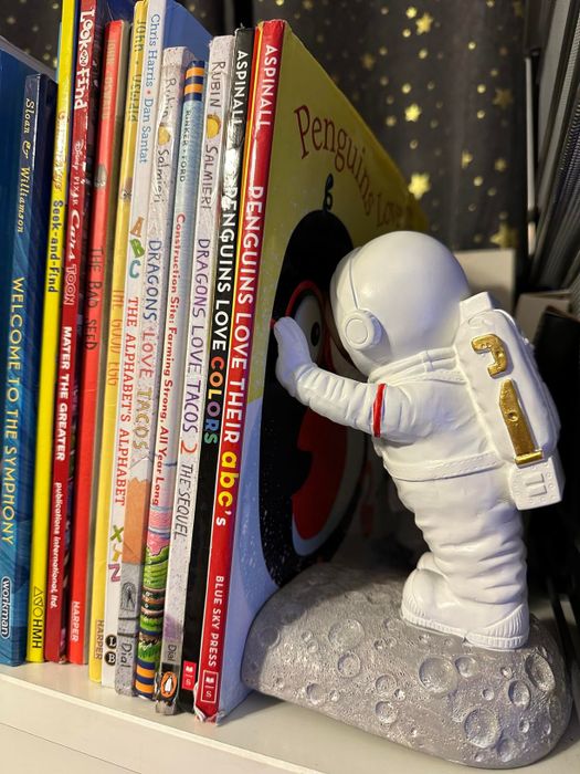 An astronaut figure bookend supporting a stack of children's books on a shelf.