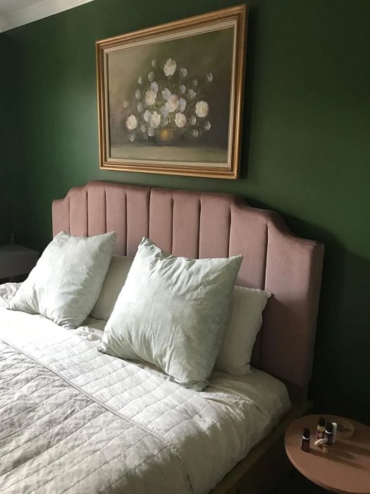 A bedroom featuring a padded headboard, comfy pillows, a quilted bedspread, a small round bedside table with nail polish, and a framed floral painting on the wall.