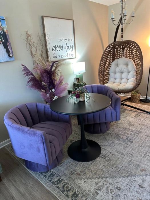 Two purple velvet chairs, a black round table adorned with a floral centerpiece, a wicker egg chair, and a wall sign reading.