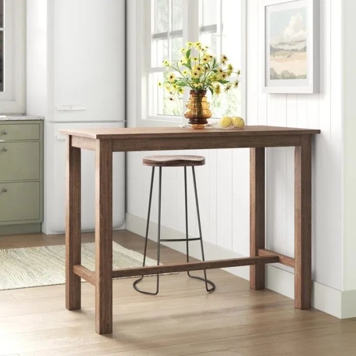 A tall wooden table with a stool placed beside it in a bright corner. The table is adorned with a vase of flowers and other decorative items.