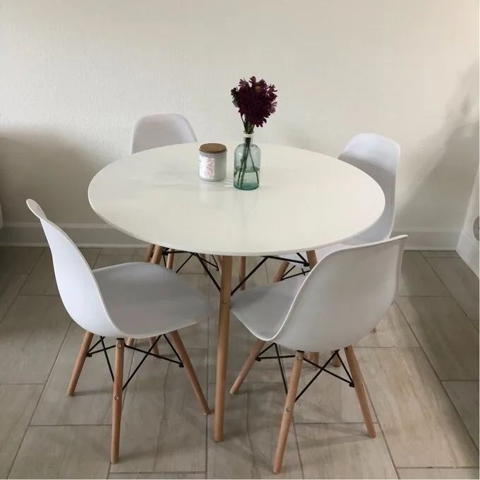 A white round dining table surrounded by four sleek modern chairs, featuring a candle and a vase with fresh flowers, set in a minimalist room.