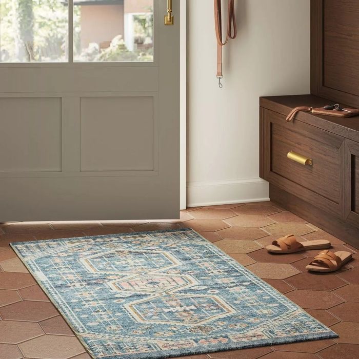 A stylish patterned rug laid out on hexagonal tiles in an entryway, beside a door, key hooks, and a shoe bench with leather sandals resting nearby.
