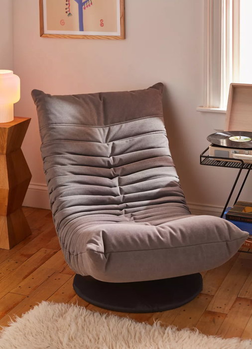 A gray upholstered low lounge chair featuring horizontal pleats and supported by a black circular base, set on a hardwood floor