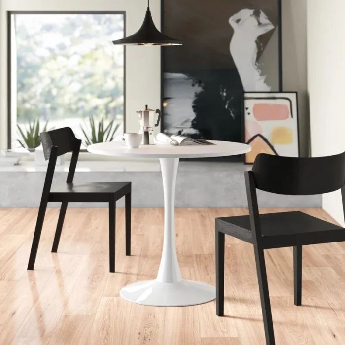 A modern dining setup featuring a white round table with a silver coffee pot and two black chairs in a minimalist room with a wooden floor and abstract wall art.