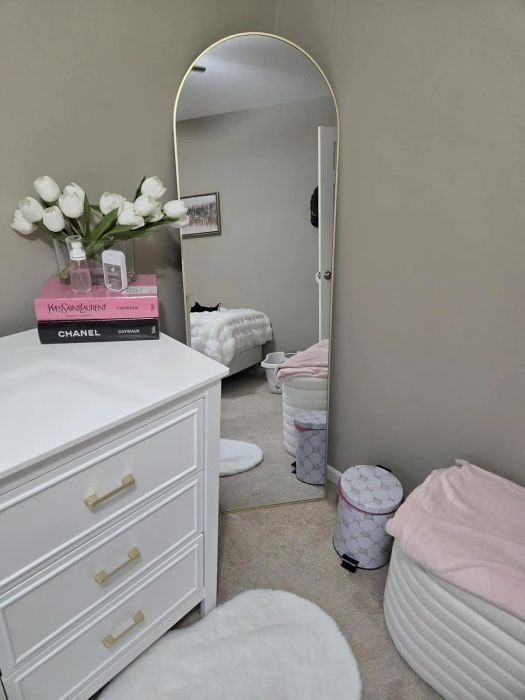 A cozy bedroom corner featuring a dresser with books and a vase of white tulips, a large arched mirror, an ottoman, and a reflection of the bed.