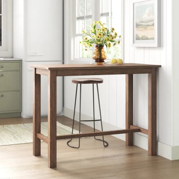 A wooden dining table with a tall stool, highlighted by a vase of bright yellow flowers and fresh lemons, set in a sun-filled kitchen.