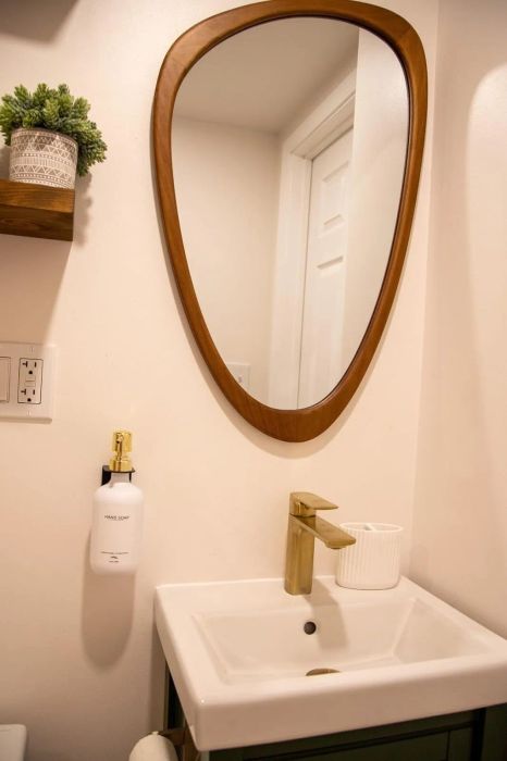 A sleek modern bathroom showcasing a white sink with a golden faucet, a rounded mirror, a soap dispenser, and a small potted plant resting on a shelf.