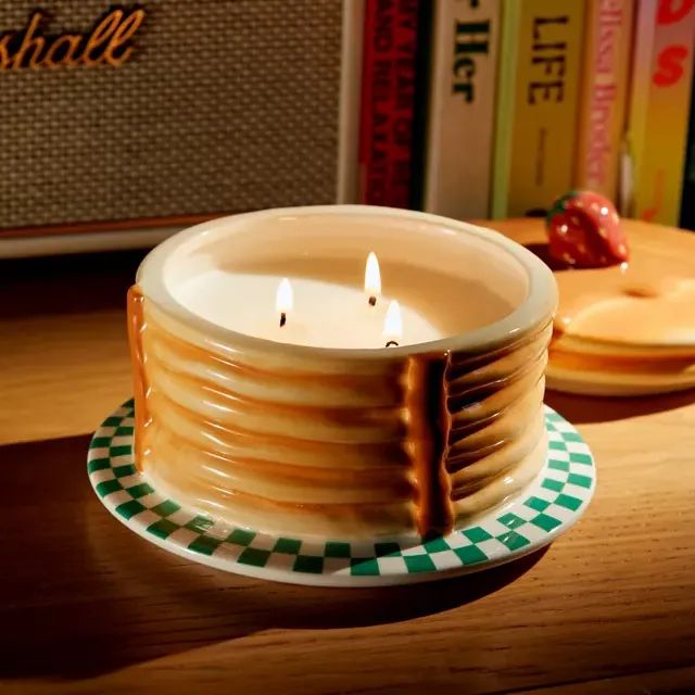 A pancake-shaped candle with three wicks resting on a checkered plate, placed on a wooden surface with books and a speaker in the background.