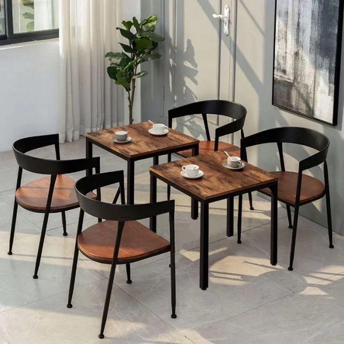 Two square wooden tables paired with four matching black-framed chairs, set in a modern, sunlit room with a potted plant and a large artwork.