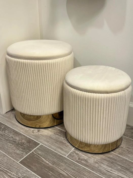 Two elegant cream-colored, pleated ottomans with gold bases are placed on a wooden-tiled floor.