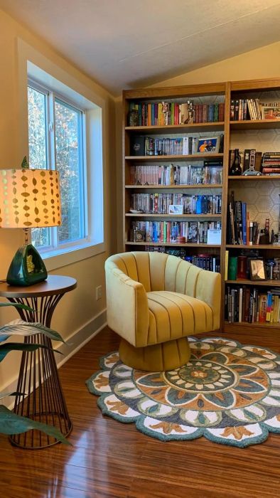 A cozy reading nook featuring a plush round chair in front of a bookcase filled with various books, DVDs, and decorative items. A side table with a lamp is nearby.