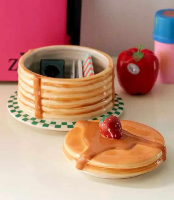 A pancake-shaped ceramic organizer with a lid, used to store small items, placed on a green checkered plate. In the background, decorative apples and skincare products can be seen.