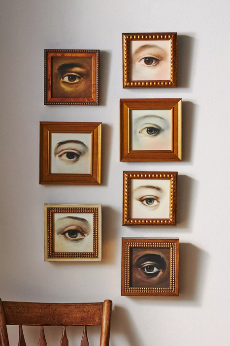 A set of six framed art pieces featuring lifelike eye illustrations arranged in a grid above a wooden chair