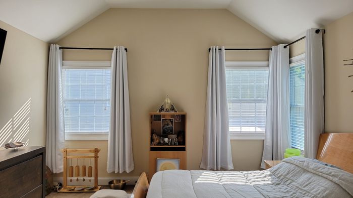 A cozy bedroom featuring a bed with a white quilt, a wooden headboard, and a dresser. Two windows with white curtains let in natural light, and a small wooden shelf adds decor to the space.