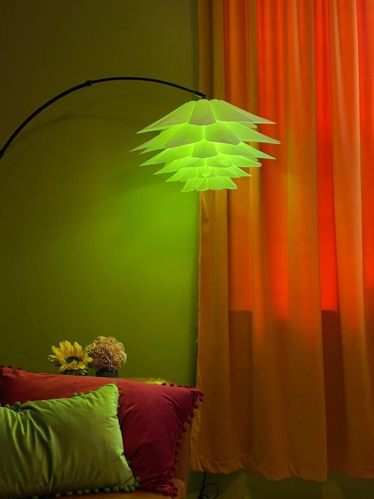 A modern geometric lamp with a sleek design hangs beside a window adorned with colorful curtains. Below, a collection of vibrant pillows rests alongside a pot of sunflowers.