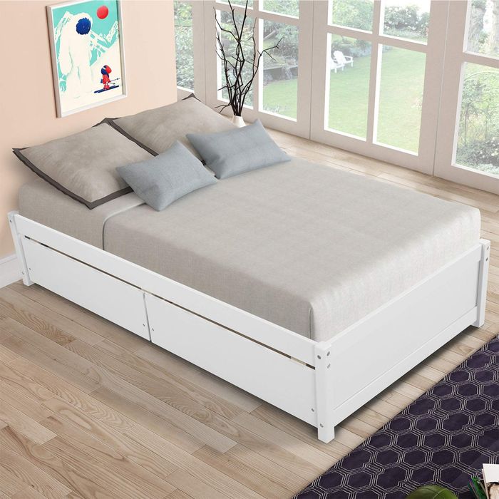 A white wooden bed with soft-colored bedding, gray pillows, and hidden trundle storage in a bright, airy room with large windows. A piece of modern artwork and a vase sit beside the bed.