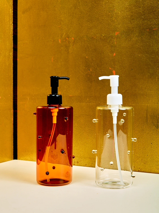 Two pump bottles sitting on a surface: one amber with a black pump and the other clear with a white pump, both featuring decorative bubble details.