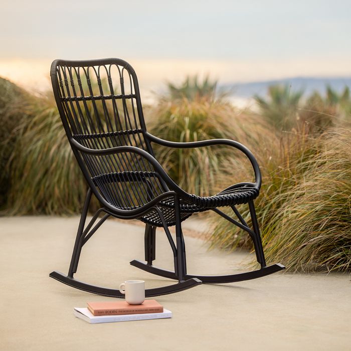 A rattan rocking chair on an outdoor patio, with a mug and a book resting on a slim platform nearby