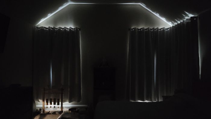 A dimly lit room with long curtains drawn shut, blocking most light from entering. A chair rests in the corner, and the edge of a bed is visible.