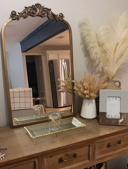 An arched mirror with a gold-tone frame, complete with elegant scrolling details, placed atop a reviewer’s vanity table