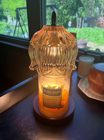 A Tuscany Candle Co. Lemon Sugar Cookie scented candle lit on a dark surface, encased in a decorative glass cover, with sunlight streaming through a window in the background.